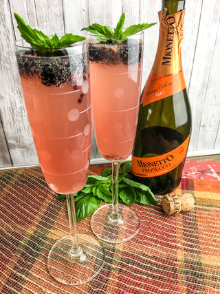 rose colored cocktail in a champagne flute with garnishes of fresh blackberry and basil