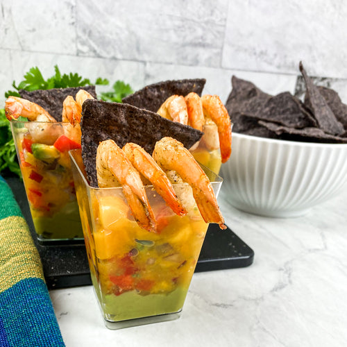 Mango Salsa Shrimp Cups recipe image with colorful layers of mango salsa, and guacamole topped with fresh shrimp with a bowl of blue corn chips in the background on a white countertop